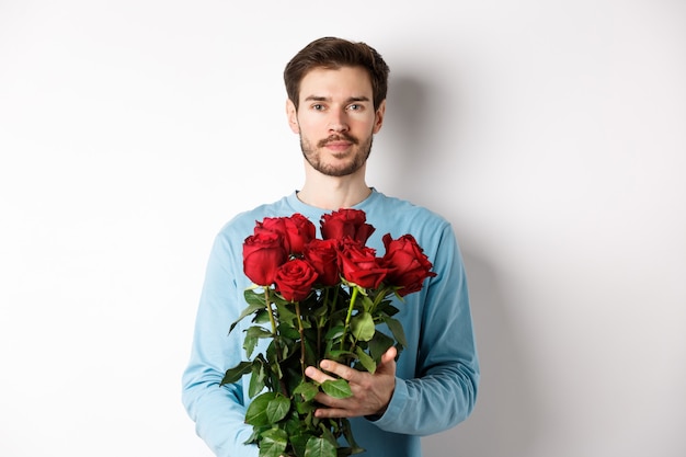 Jovem confiante trazendo flores no dia dos namorados, segurando um buquê romântico, de pé sobre um fundo branco