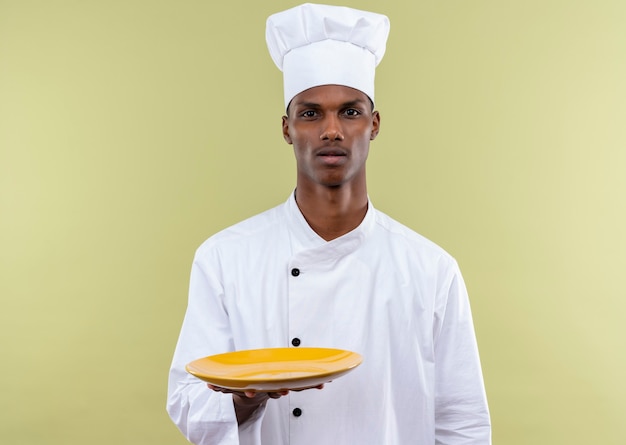 Jovem confusa cozinheira afro-americana com uniforme de chef segurando um prato isolado na parede verde