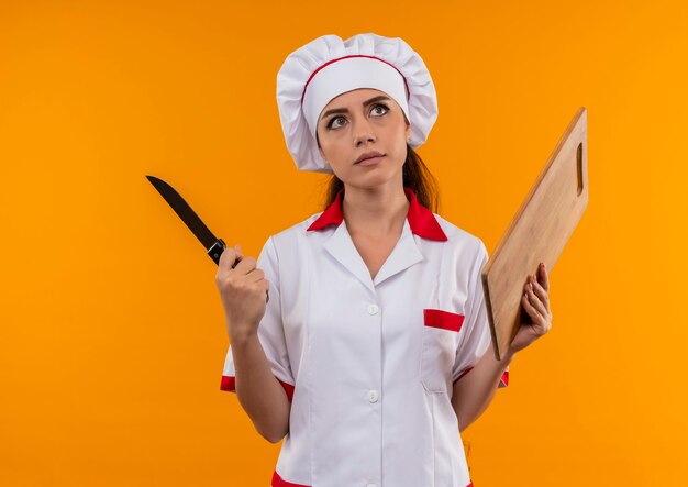 Jovem confusa cozinheira caucasiana com uniforme de chef segurando uma faca e uma tábua de madeira isoladas na parede laranja com espaço de cópia