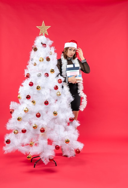 Jovem confusa e linda mulher com chapéu de Papai Noel em pé atrás de uma árvore de Natal decorada segurando presentes e procurando algo