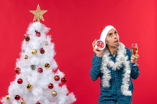 Jovem confuso com chapéu de Papai Noel, segurando uma taça de vinho e um relógio perto da árvore de Natal