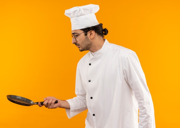 Jovem confuso cozinheiro usando uniforme de chef e óculos segurando e olhando para uma frigideira