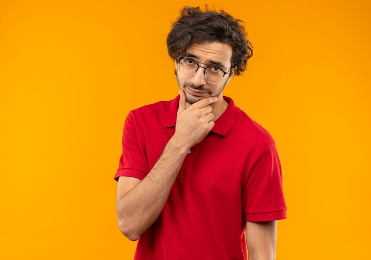 Foto grátis jovem confuso de camisa vermelha com óculos ópticos coloca a mão no queixo e parece isolado na parede laranja