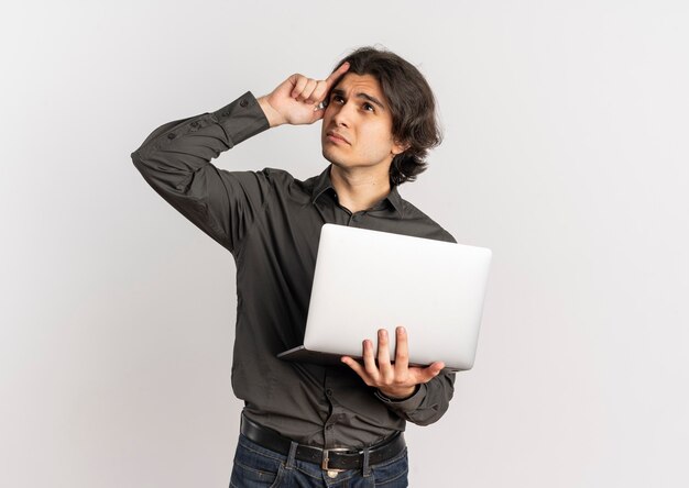 Jovem confuso e bonito homem caucasiano coloca a mão na cabeça e segura o laptop isolado no fundo branco com espaço de cópia