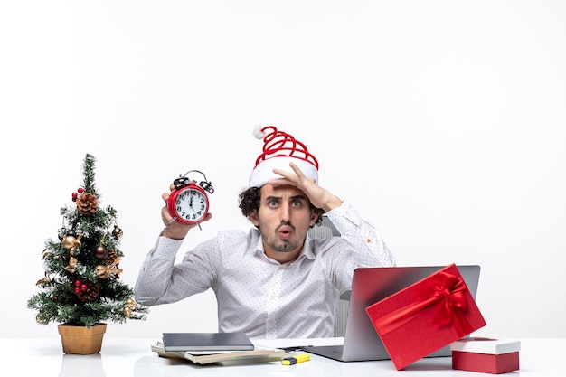 Jovem confuso empresário com chapéu de papai noel e mostrando o relógio e brainstorming sentado no escritório sobre fundo branco