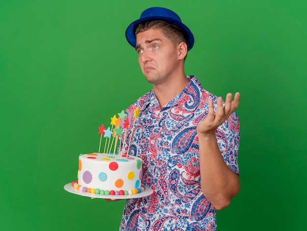 Jovem confuso festeiro olhando para o lado com chapéu azul, segurando o bolo e estendendo a mão isolada no verde