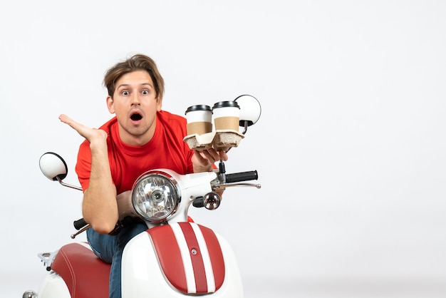 Foto grátis jovem confuso mensageiro de uniforme vermelho sentado na scooter segurando pedidos na parede amarela