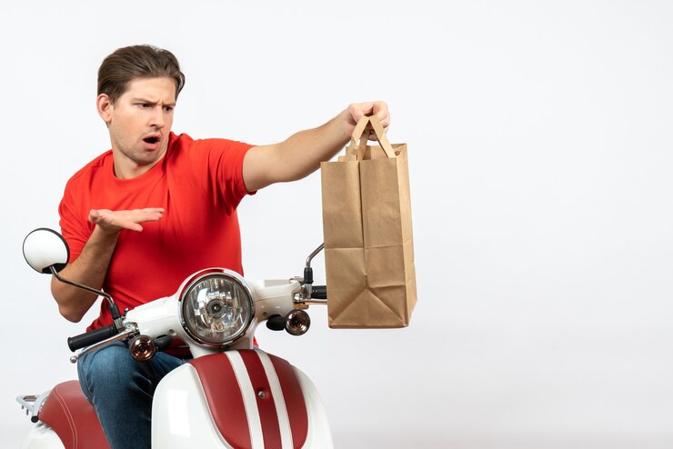 Foto grátis jovem confuso mensageiro emocional em uniforme vermelho sentado na scooter segurando um saco de papel na parede branca