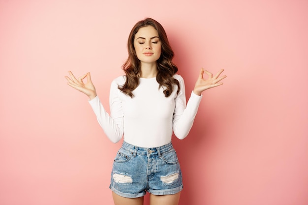 Foto grátis jovem consciente e relaxada meditando, mantenha a calma, relaxando durante a ioga, feche os olhos e dobre os dedos em gesto de mudra, fundo rosa