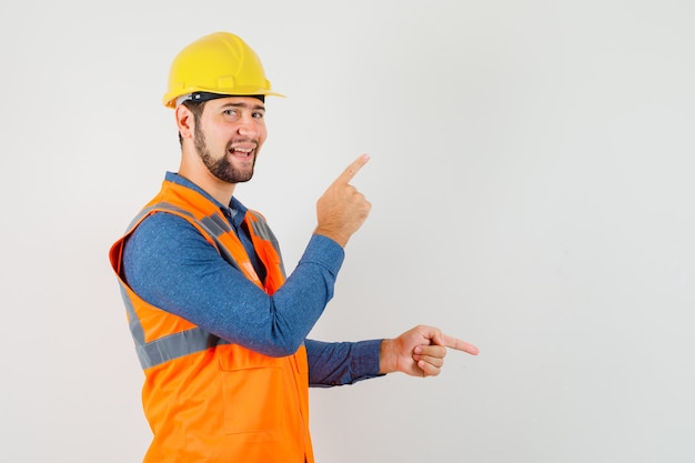 Jovem construtor apontando os dedos para cima e para baixo na camisa, colete, capacete e parecendo alegre. .