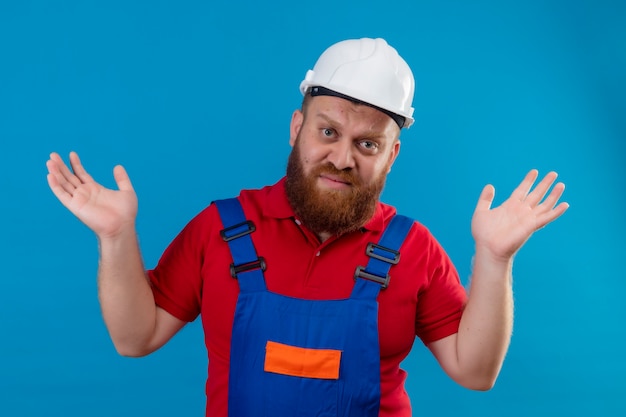 Jovem construtor barbudo em uniforme de construção e capacete de segurança encolhendo os ombros, parecendo confuso e inseguro, sem resposta, espalhando as palmas das mãos