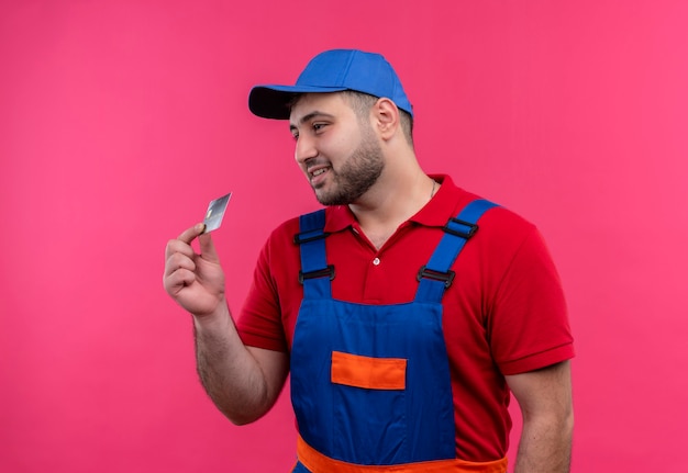 Foto grátis jovem construtor com uniforme de construção e boné mostrando cartão de crédito olhando para o lado sorrindo