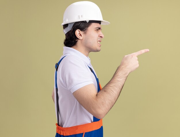 Jovem construtor com uniforme de construção e capacete de segurança apontando com o dedo indicador para o lado, sendo confundido em pé de lado sobre a parede verde