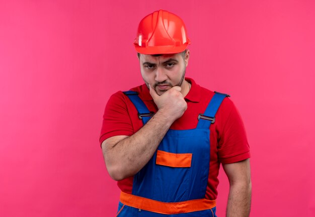 Jovem construtor com uniforme de construção e capacete de segurança olhando para a câmera com o rosto carrancudo e a mão no queixo com expressão pensativa