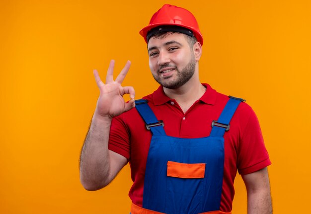 Foto grátis jovem construtor com uniforme de construção e capacete de segurança olhando para a câmera sorrindo alegremente fazendo sinal de ok