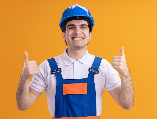 Foto grátis jovem construtor com uniforme de construção e capacete de segurança olhando para a frente, sorrindo alegremente, mostrando os polegares em pé sobre a parede laranja