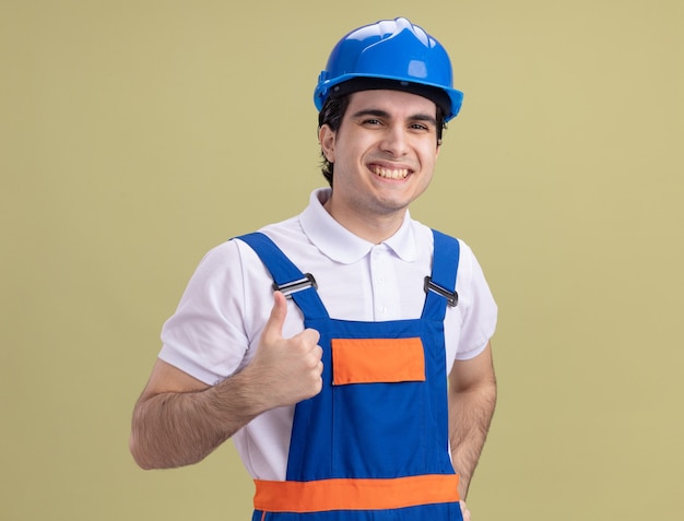 Jovem construtor com uniforme de construção e capacete de segurança olhando para a frente sorrindo alegremente mostrando os polegares em pé sobre a parede verde