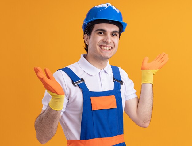 Jovem construtor com uniforme de construção e capacete de segurança, olhando para a frente, sorrindo com uma cara feliz em pé sobre a parede laranja