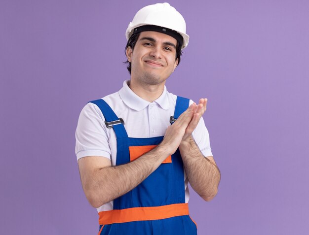 Jovem construtor com uniforme de construção e capacete de segurança, olhando para a frente, sorrindo confiante, aplaudindo em pé sobre a parede roxa