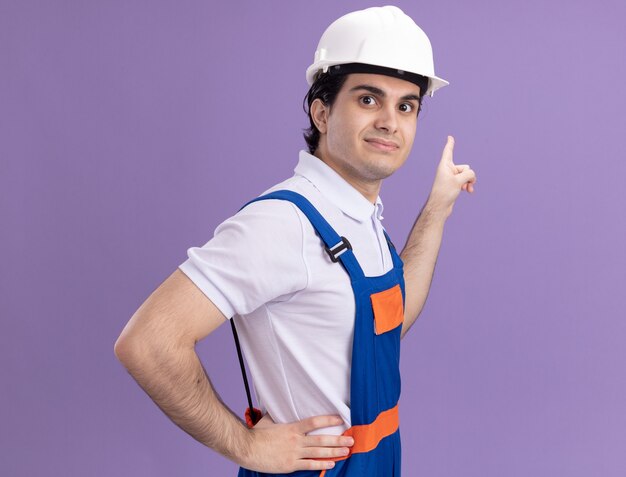 Jovem construtor com uniforme de construção e capacete de segurança, olhando para a frente, sorrindo confiante, apontando com o dedo indicador para algo em pé sobre a parede roxa