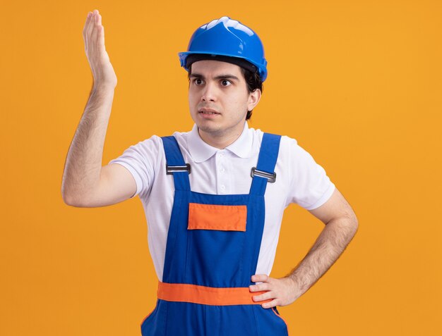 Jovem construtor com uniforme de construção e capacete de segurança, olhando para o lado, confuso, levantando a mão em desprazer em pé sobre a parede laranja