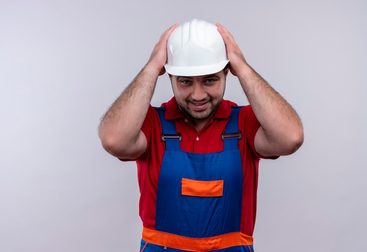 Foto grátis jovem construtor com uniforme de construção e capacete de segurança, parecendo desapontado e confuso com o toque na cabeça