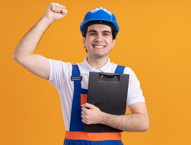 Jovem construtor com uniforme de construção e capacete de segurança segurando a prancheta, olhando para a frente, feliz e positivo levantando o punho em pé sobre a parede laranja