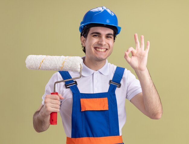 Jovem construtor com uniforme de construção e capacete de segurança segurando o rolo de pintura, olhando para frente com um sorriso no rosto, mostrando uma placa de ok em cima da parede verde