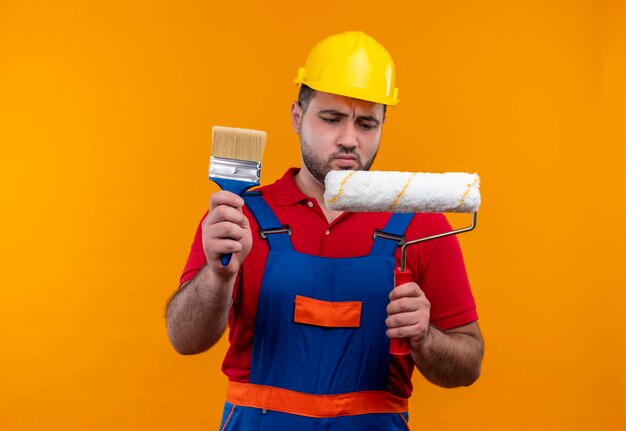 Jovem construtor com uniforme de construção e capacete de segurança segurando rolo de pintura e pincel olhando para rolo de pintura com expressão cética