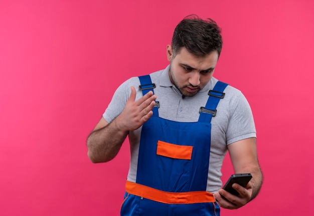 Jovem construtor com uniforme de construção segurando um smartphone, olhando para a tela descontente e confuso