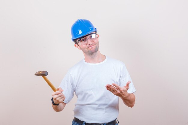 Jovem construtor de camiseta, jeans, capacete mostrando o martelo e parecendo desapontado