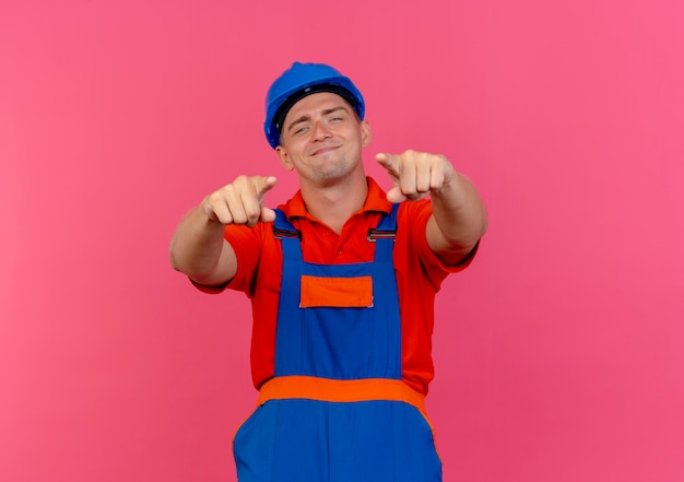 Foto grátis jovem construtor do sexo masculino satisfeito usando uniforme e capacete de segurança apontando para a câmera na cor rosa