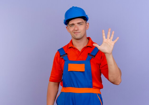 Jovem construtor do sexo masculino satisfeito usando uniforme e capacete de segurança mostrando cinco em roxo