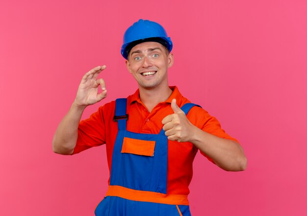 Foto grátis jovem construtor do sexo masculino satisfeito usando uniforme e capacete de segurança, mostrando okey gesto com o polegar para cima na rosa