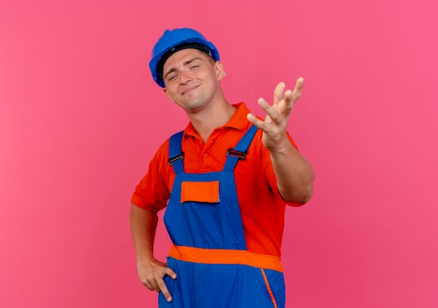 Jovem construtor do sexo masculino satisfeito, vestindo uniforme e capacete de segurança, estendendo a mão para a câmera e colocando a outra mão no quadril rosa