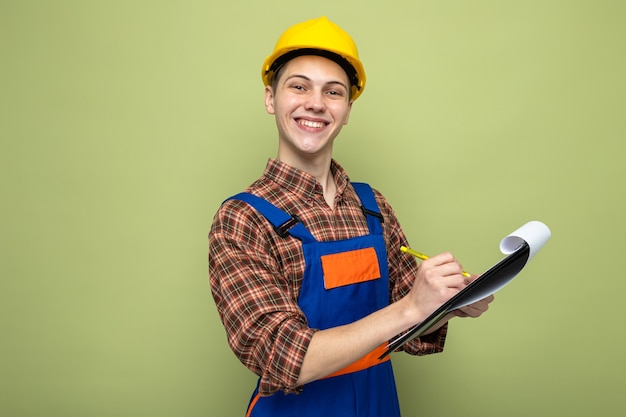 Jovem construtor do sexo masculino sorridente escreve na prancheta usando uniforme