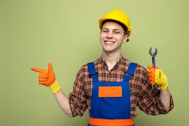 Jovem construtor do sexo masculino sorrindo olhando de lado usando uniforme com luvas segurando uma chave inglesa