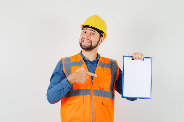 Jovem construtor em camisa, colete, capacete, apontando para a área de transferência e olhando alegre, vista frontal.