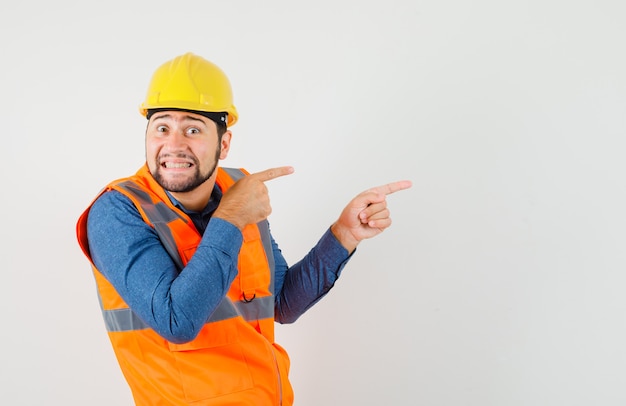 Jovem construtor em camisa, colete, capacete apontando para o lado e parecendo curioso, vista frontal.