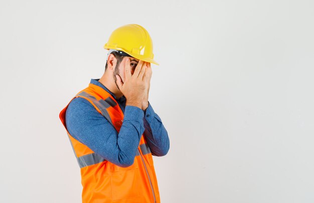 Jovem construtor em camisa, colete, capacete, olhando por entre os dedos e parecendo assustado, vista frontal.