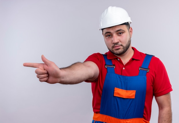 Jovem construtor em uniforme de construção e capacete de segurança apontando com o dedo indicador para os lados com uma cara séria