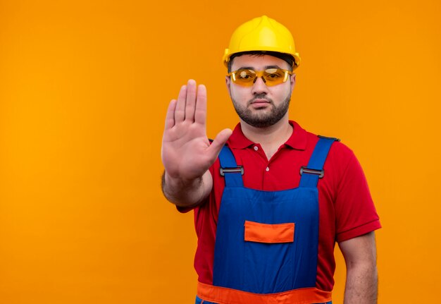 Jovem construtor em uniforme de construção e capacete de segurança com a mão aberta, fazendo sinal de pare com uma cara séria