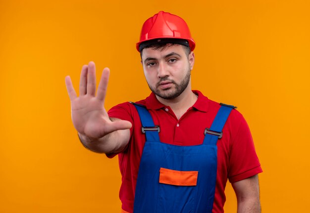 Jovem construtor em uniforme de construção e capacete de segurança com a mão aberta, fazendo sinal de pare com uma cara séria