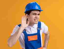 Foto grátis jovem construtor em uniforme de construção e capacete de segurança olhando para frente com a mão na orelha tentando ouvir fofocas sendo confundidas em pé sobre a parede laranja