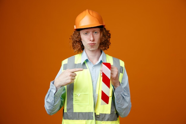 Jovem construtor em uniforme de construção e capacete de segurança segurando fita adesiva apontando com o dedo indicador para ele parecendo confiante em pé sobre fundo laranja