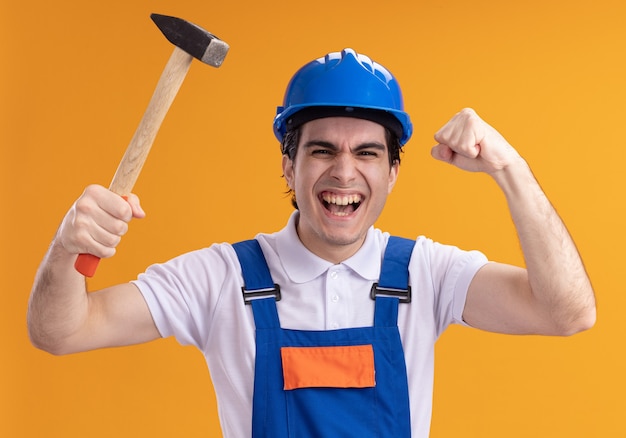 Jovem construtor em uniforme de construção e capacete de segurança segurando um martelo, olhando para a frente, feliz e animado, punho cerrado em pé sobre a parede laranja