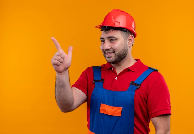 Jovem construtor em uniforme de construção e capacete de segurança sorrindo alegremente apontando com o dedo indicador para o lado