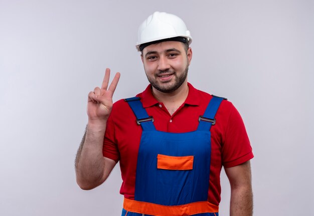 Jovem construtor em uniforme de construção e capacete de segurança sorrindo, mostrando e apontando para cima com os dedos número dois