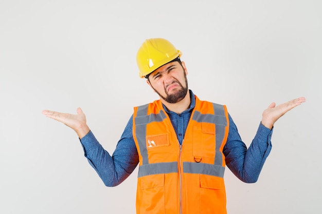 Jovem construtor fazendo gesto de escalas enquanto carranca na camisa, colete, vista frontal do capacete.