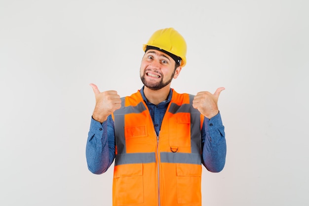 Jovem construtor mostrando dois polegares para cima na camisa, colete, capacete e olhando alegre, vista frontal.